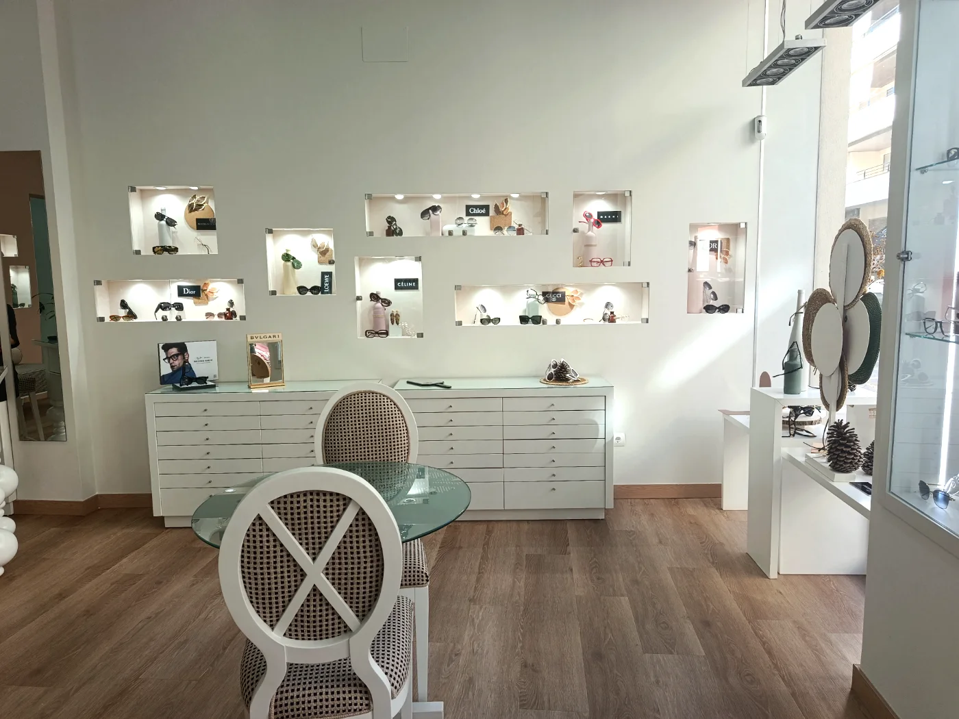 Zona de atención de Senti2 Óptica: mesa de cristal, sillas, cajoneras blancas y vitrinas de marcas